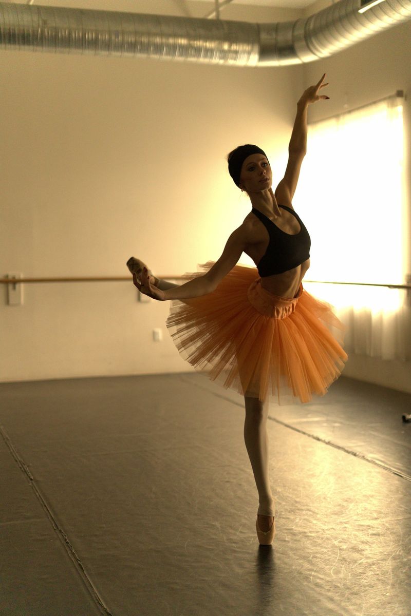 Dancer in an orange tutu on pointe with one arm raised in a sunlit studio